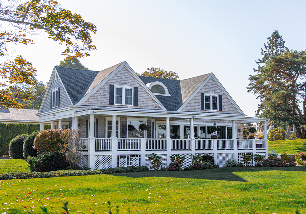 A large white house with a wrap around porch on a sunny day in front of trees with a beautiful lawn.