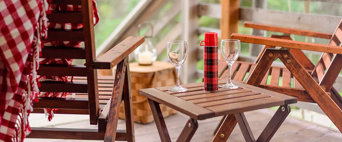An outside close up of Two wine glasses and a plaid Thermos sitting on a wooden table next to two chairs